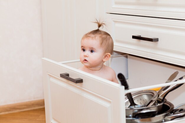 Joyeux enfant assis dans le tiroir de la cuisine avec des casseroles et riant. Portrait d'un enfant dans une cuisine blanche.