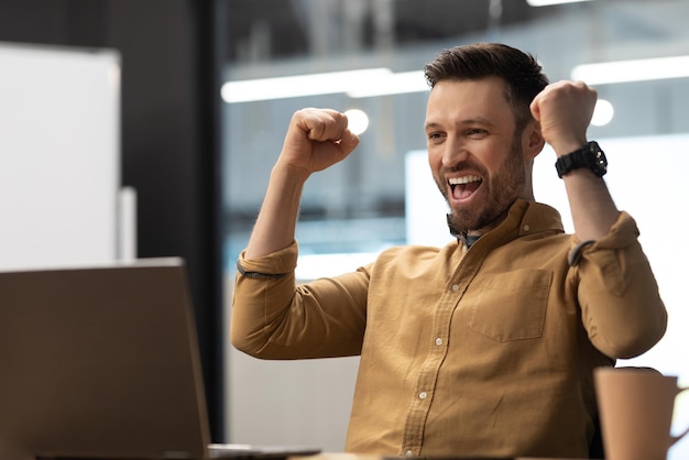 Joyeux entrepreneur masculin regardant un ordinateur portable secouant les poings au bureau