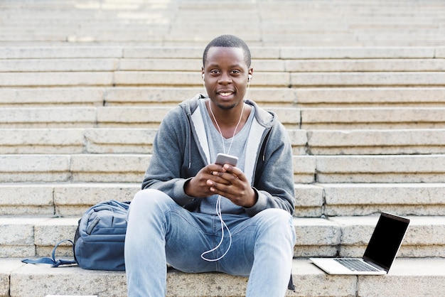 Joyeux étudiant afro-américain assis dans les escaliers et écoutant de la musique sur son smartphone à l'extérieur, se reposant sur le campus universitaire avec espace de copie. Concept d'éducation, espace de copie