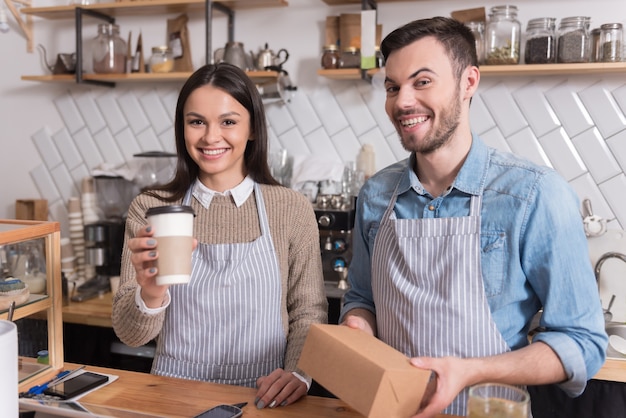 Joyeux festin. Agréable jeune couple tenant la boîte et une tasse de café et souriant tout en se tenant derrière le bar.