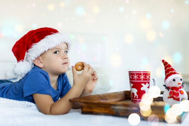 Joyeux garçon au lit mangeant des biscuits et buvant du cacao Dans le chapeau du père Noël Bokeh sur fond Noël