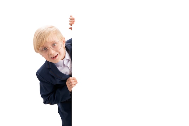 Joyeux garçon blond dans de grands verres et uniforme scolaire jetant un coup d'œil derrière un fond blanc Copier l'espace Studio shot Isoler