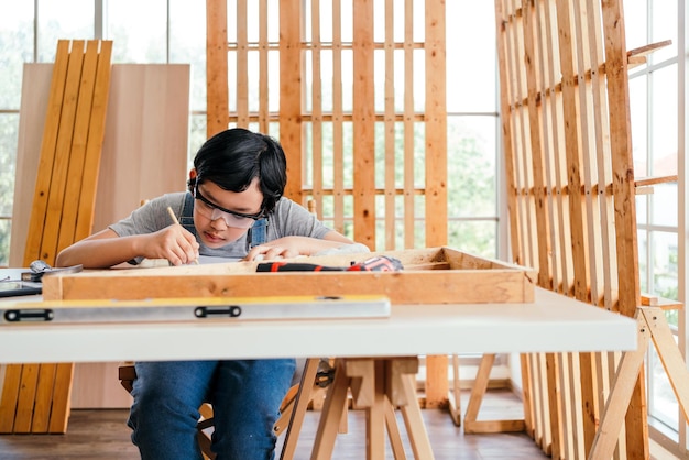 Joyeux garçon charpentier asiatique portant des lunettes de sécurité et des gants de travail dessinant délibérément un modèle de construction sur la table Travaux de construction et de réparation Concept de charpentier et de famille