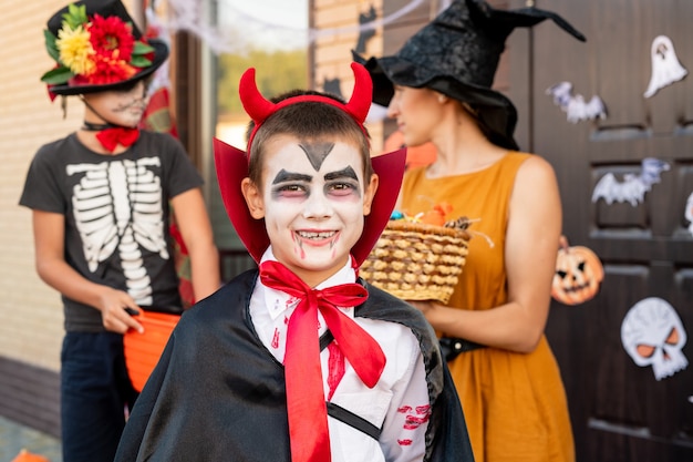 Joyeux garçon en costume d'halloween vous regarde contre jeune femme en robe jaune et chapeau tenant un panier avec des friandises et son ami