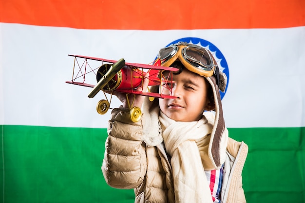 Joyeux garçon indien ou asiatique jouant avec un avion en métal jouet dans une tenue et des lunettes de pilote de la seconde guerre mondiale, debout isolé sur fond de drapeau indien