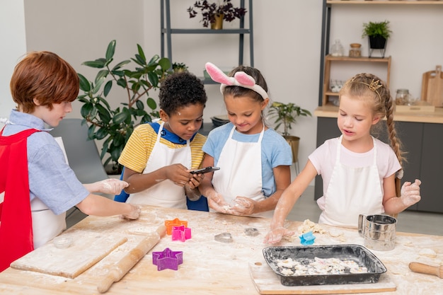 Joyeux garçon mignon avec des biscuits faits maison crus vous regardant en vous tenant debout près de la table de la cuisine