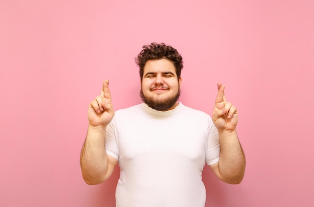 Joyeux garçon en surpoids croisa les doigts les yeux fermés portant un t-shirt blanc isolé