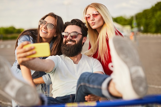 Joyeux gars ethnique assis dans une charrette à bras et prenant un selfie avec des amies