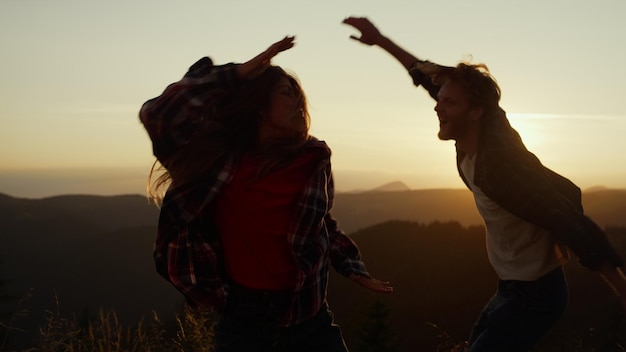 Joyeux gars et fille sautant dans l'air en plein air Homme et femme positifs dansant dans les montagnes au coucher du soleil Couple souriant gesticulant les mains pendant la danse Hipsters féminins et masculins passant du temps ensemble