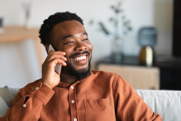 Joyeux gars noir parlant sur un téléphone portable assis sur un canapé à l'intérieur