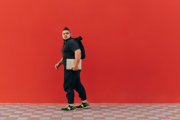 Joyeux gros homme en vêtements noirs élégants marche sur le fond d'un mur rouge avec un ordinateur portable