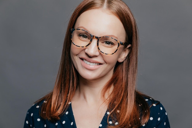 Joyeux gros plan d'une jolie femme aux cheveux bruns, le sourire à pleines dents porte des lunettes habillées en chemisier formel pose sur fond gris, heureuse de rencontrer des collègues lors d'une réunion