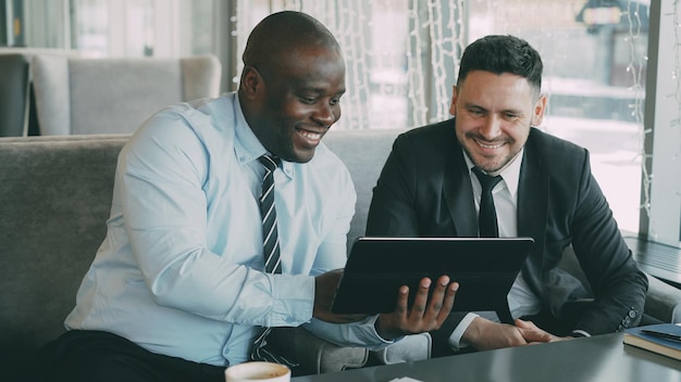 Joyeux homme d'affaires afro-américain en vêtements formels à l'aide d'une tablette numérique discutant d'un projet de démarrage avec son partenaire caucasien dans un café élégant pendant la pause déjeuner