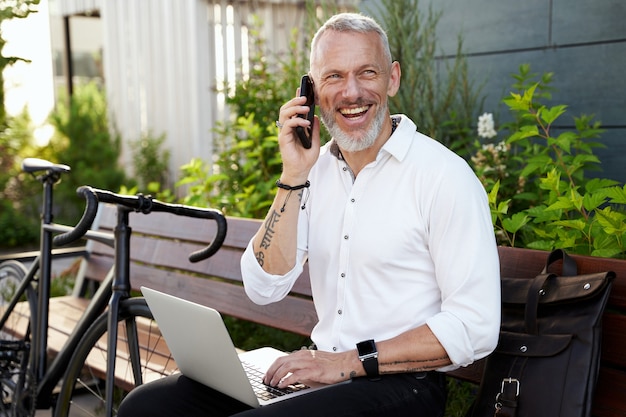 Joyeux homme d'affaires d'âge moyen souriant parlant au téléphone à l'aide de son ordinateur portable assis sur le