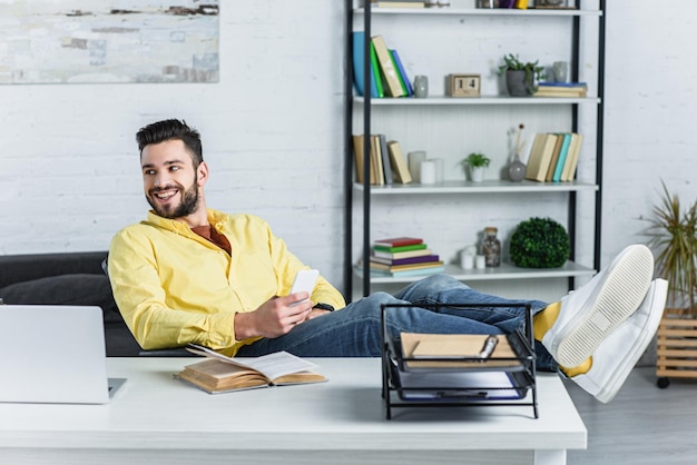 Joyeux homme d'affaires barbu tenant un smartphone et regardant loin sur le lieu de travail