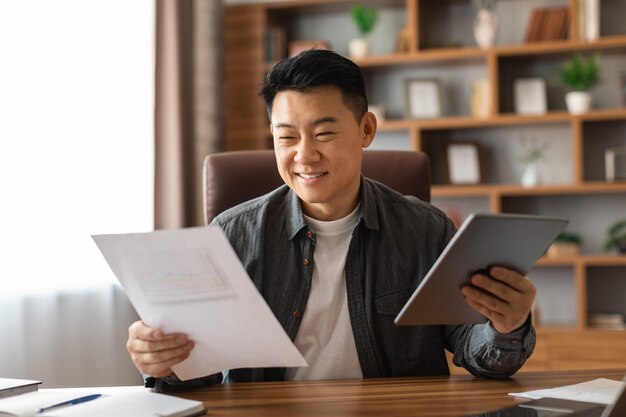 Joyeux homme d'affaires japonais d'âge moyen avec des documents et une tablette travaille à l'intérieur du bureau à domicile