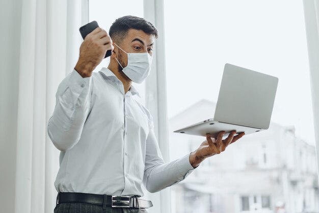 Joyeux homme d'affaires en masque médical tenant un ordinateur portable et un téléphone portable célèbre le succès de près