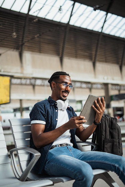 Joyeux homme africain utilisant une tablette alors qu'il est assis sur un banc dans une gare