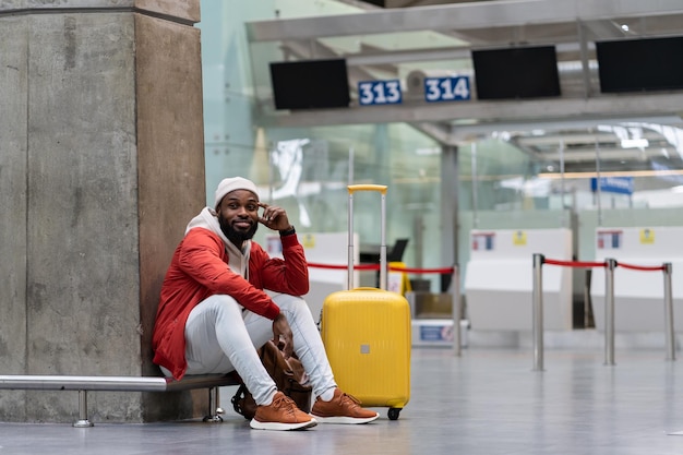 Joyeux homme afro-américain assis dans le terminal de l'aéroport avec une valise jaune près des comptoirs d'enregistrement