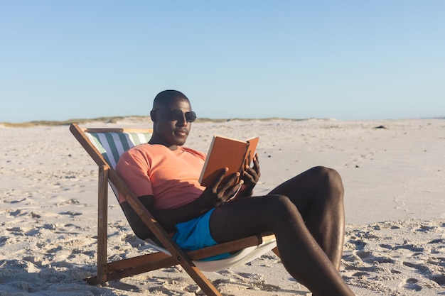 Photo joyeux homme afro-américain en lunettes de soleil assis dans un transat lisant un livre sur une plage ensoleillée. été, détente, temps libre et vacances, inchangés.