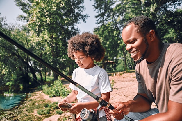 Joyeux homme afro-américain et son adorable fils qui ont l'air concentrés tout en tenant des cannes à pêche