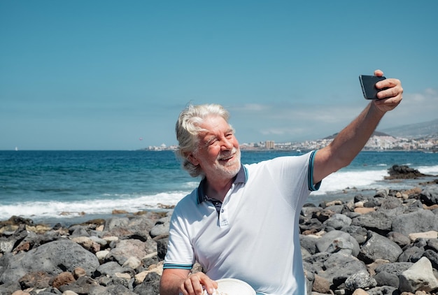 Joyeux homme âgé aux cheveux blancs profitant de vacances à la mer, assis à la plage par une journée ensoleillée, prenant un selfie avec un téléphone portable