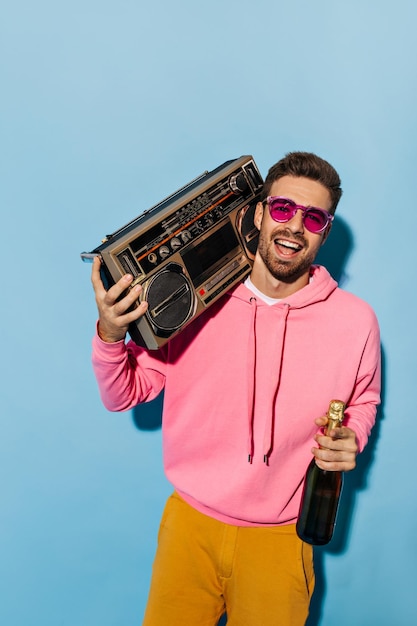 Joyeux homme barbu joyeux en short jaune et sweat à capuche rose sourit tient une bouteille de champagne et un tourne-disque sur fond bleu