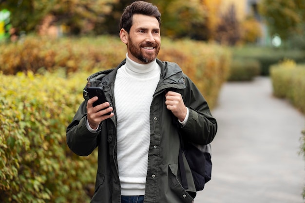 Joyeux homme brune avec une barbe à l'aide d'un téléphone portable et souriant Un gars heureux avec un sac à dos marche dans une rue de la ville