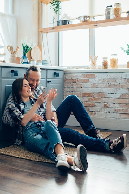 Joyeux homme et femme allongé sur le sol de la cuisine et regardant l'écran du smartphone