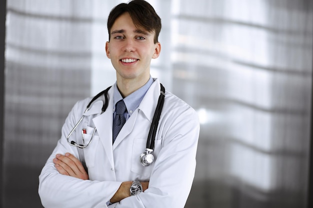 Joyeux homme-médecin souriant debout avec les bras croisés en clinique. Service médical parfait avec un jeune médecin intelligent à l'hôpital. Notion de médecine.