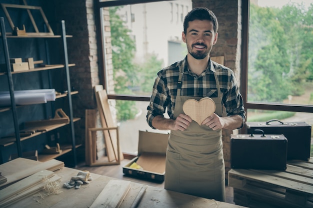 Joyeux homme séduisant positif tenant un coeur en bois fait par lui-même démontrant son talent de menuiserie