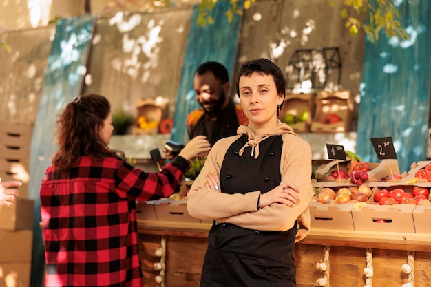 Joyeux jeune agriculteur posant à côté de l'étal du marché des agriculteurs, présentant des produits naturels sains à l'extérieur. Femme confiante vendant des produits biologiques de saison, des fruits et des légumes.