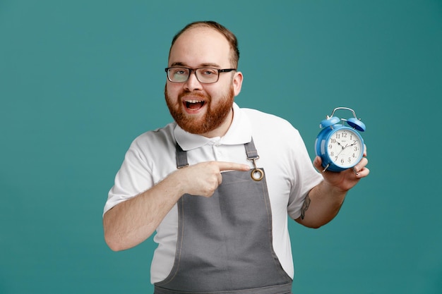 Joyeux jeune barbier portant l'uniforme et des lunettes regardant la caméra montrant un réveil pointant dessus isolé sur fond bleu