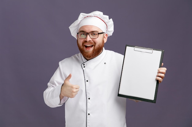 Joyeux jeune chef portant des lunettes uniformes et une casquette regardant la caméra montrant le presse-papiers et le pouce vers le haut isolé sur fond violet