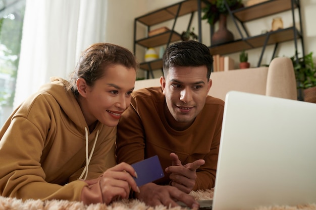 Joyeux jeune couple allongé sur le sol et achetant de nouveaux vêtements et gadgets en ligne