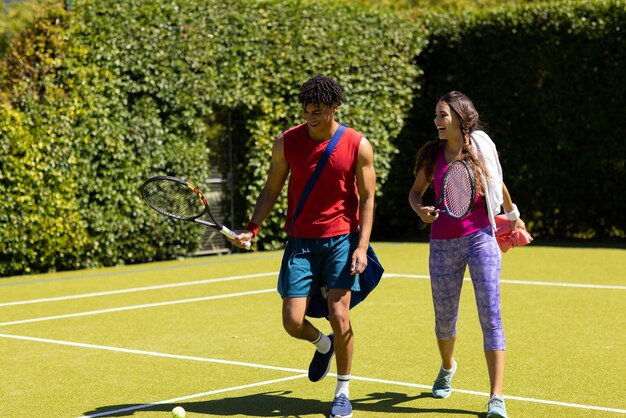 Joyeux jeune couple biracial avec des raquettes sortant du court de tennis après avoir joué au jeu. Inaltéré, amour, convivialité, sport, bonheur, compétition, passe-temps, vert, nature et concept d'été.