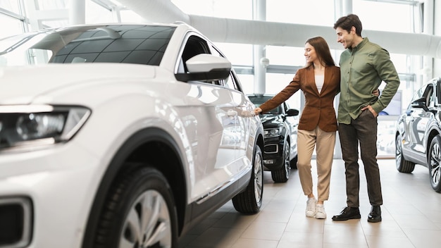 Joyeux jeune couple caucasien vérifiant la nouvelle voiture de luxe au panorama du salon automobile du concessionnaire