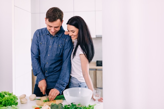 Joyeux jeune couple de famille préparant le dîner coupant une salade de légumes frais s'amusant ensemble dans un intérieur de cuisine moderne, un mari et une femme romantique heureux se liant en riant en aidant à préparer un repas sain