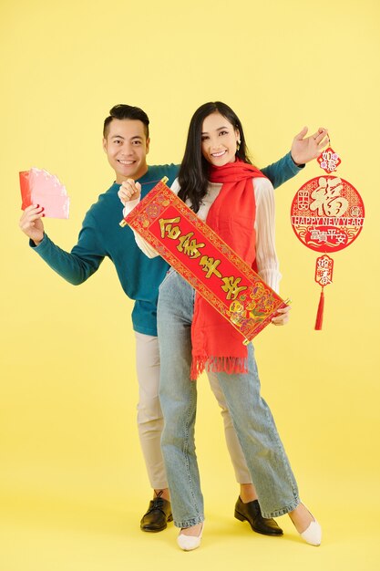 Joyeux jeune couple vietnamien en écharpes et pulls d'hiver posant avec des décorations murales avec inscription Happy New Year