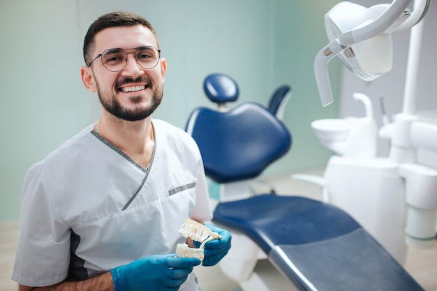 Joyeux jeune dentiste attrayant s'asseoir à l'intérieur dans la salle de dentisterie et pose