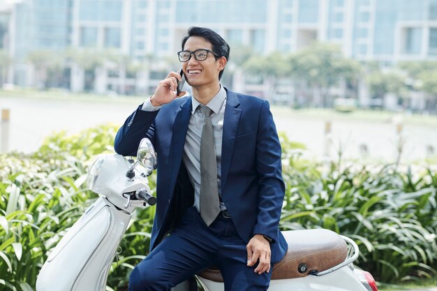 Joyeux jeune entrepreneur vietnamien assis sur un scooter et appelant au téléphone