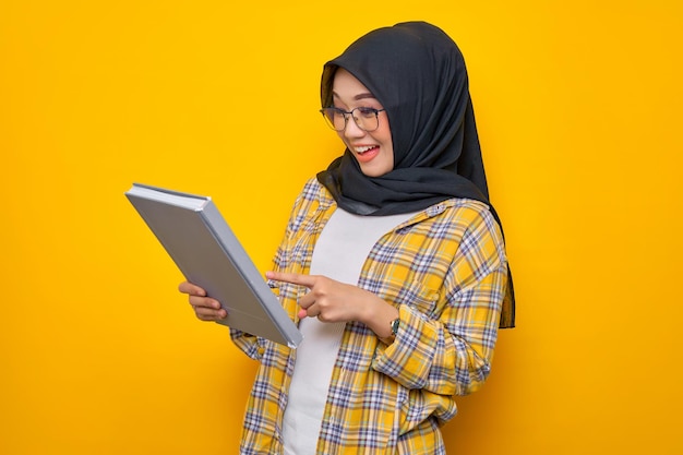 Joyeux jeune étudiant musulman asiatique tenant une liste de tâches de lecture de livre sur un livre isolé sur fond jaune Retour à l'école dans le concept de lycée