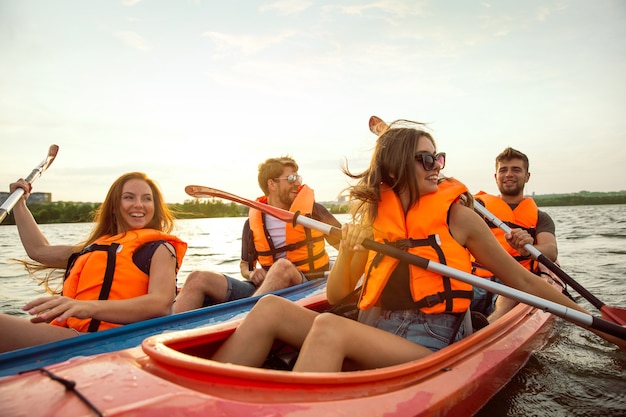Joyeux jeune groupe d'amis caucasiens faisant du kayak sur la rivière avec coucher de soleil en arrière-plan. S'amuser en loisir. Heureux modèle masculin et féminin riant sur le kayak. Sport, notion de relations.