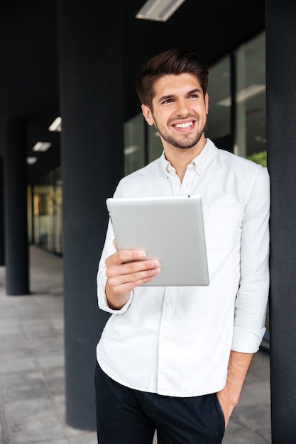 Joyeux jeune homme d'affaires debout et utilisant une tablette à l'extérieur