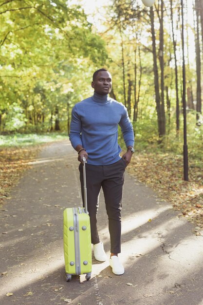 Joyeux jeune homme afro-américain dans des vêtements élégants avec une valise se promène dans un parc de printemps au chaud