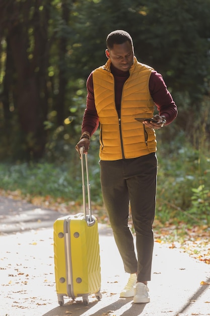 Joyeux jeune homme afro-américain dans des vêtements élégants avec une valise se promène dans un parc de printemps par une chaude journée ensoleillée Concept de voyage à la campagne