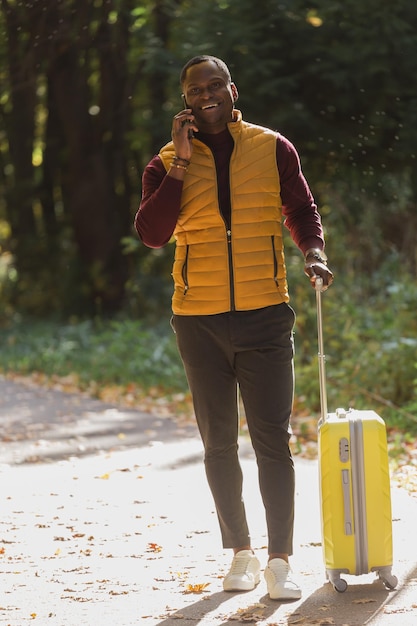 Joyeux jeune homme afro-américain dans des vêtements élégants avec une valise se promène dans un parc de printemps par une chaude journée ensoleillée Concept de voyage à la campagne