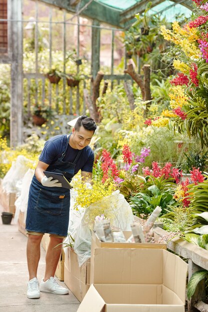 Joyeux jeune homme asiatique appréciant de travailler dans une pépinière, il déballe des boîtes de fleurs et vérifie la documentation sur une tablette