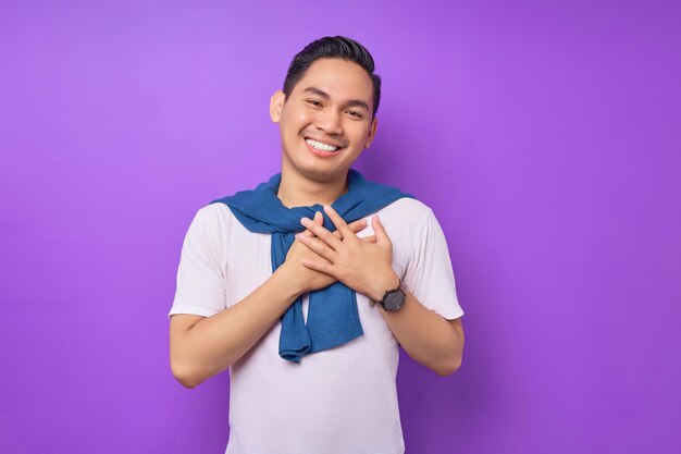 Joyeux jeune homme asiatique en colère portant un t-shirt blanc a mis sa main sur sa poitrine isolé sur fond violet concept de style de vie des gens