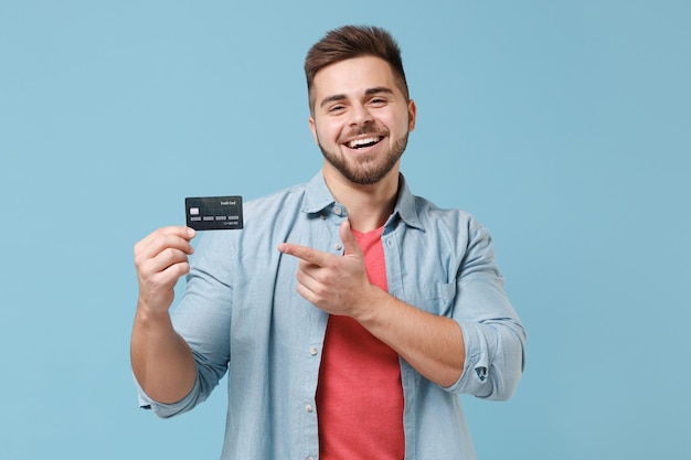 Joyeux jeune homme barbu des années 20 en chemise décontractée posant isolé sur un portrait en studio de fond de mur bleu pastel. Concept de style de vie des gens. Maquette de l'espace de copie. Pointer l'index sur la carte bancaire de crédit.